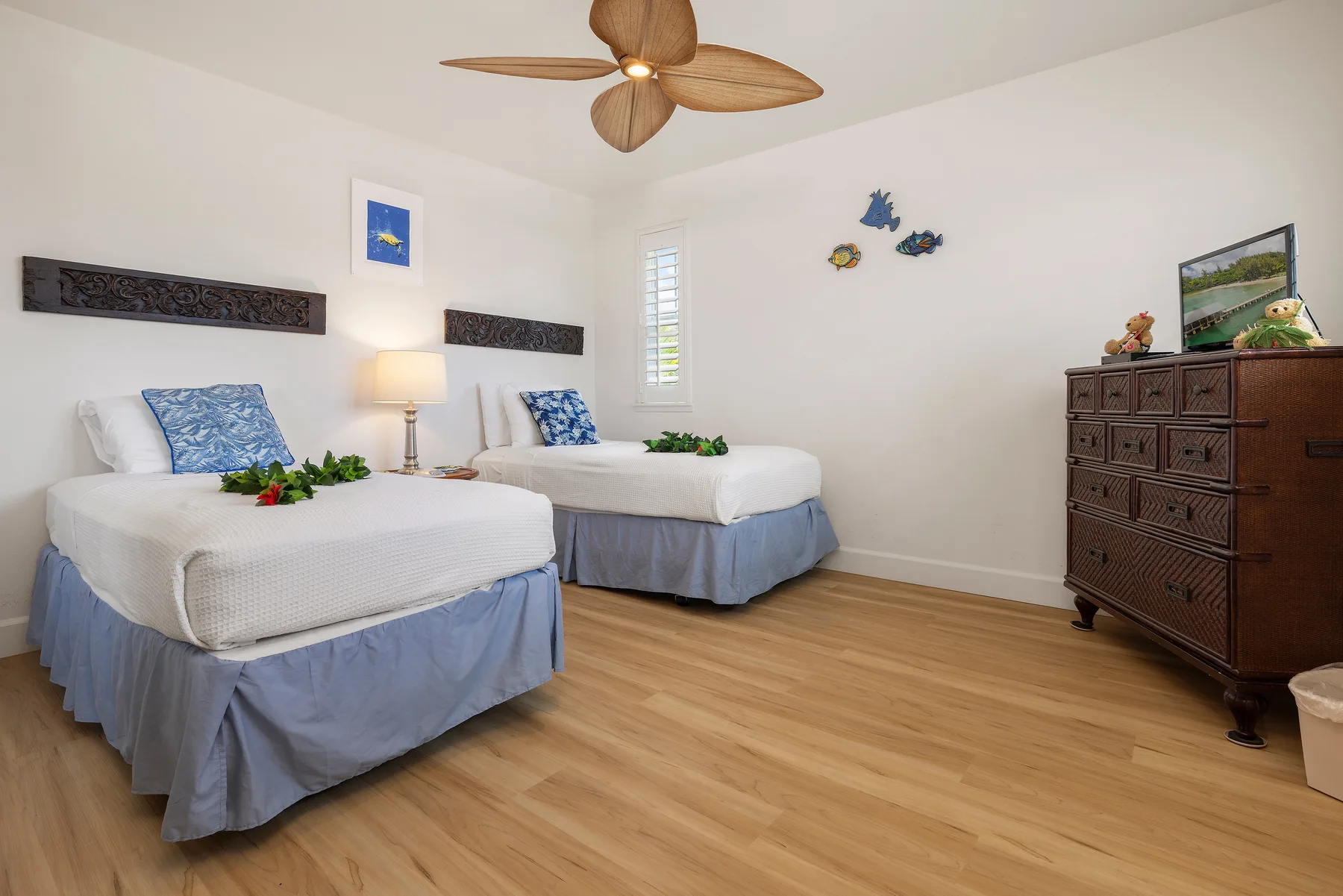 A bright bedroom has two beds with blue bedding and a wooden dresser.