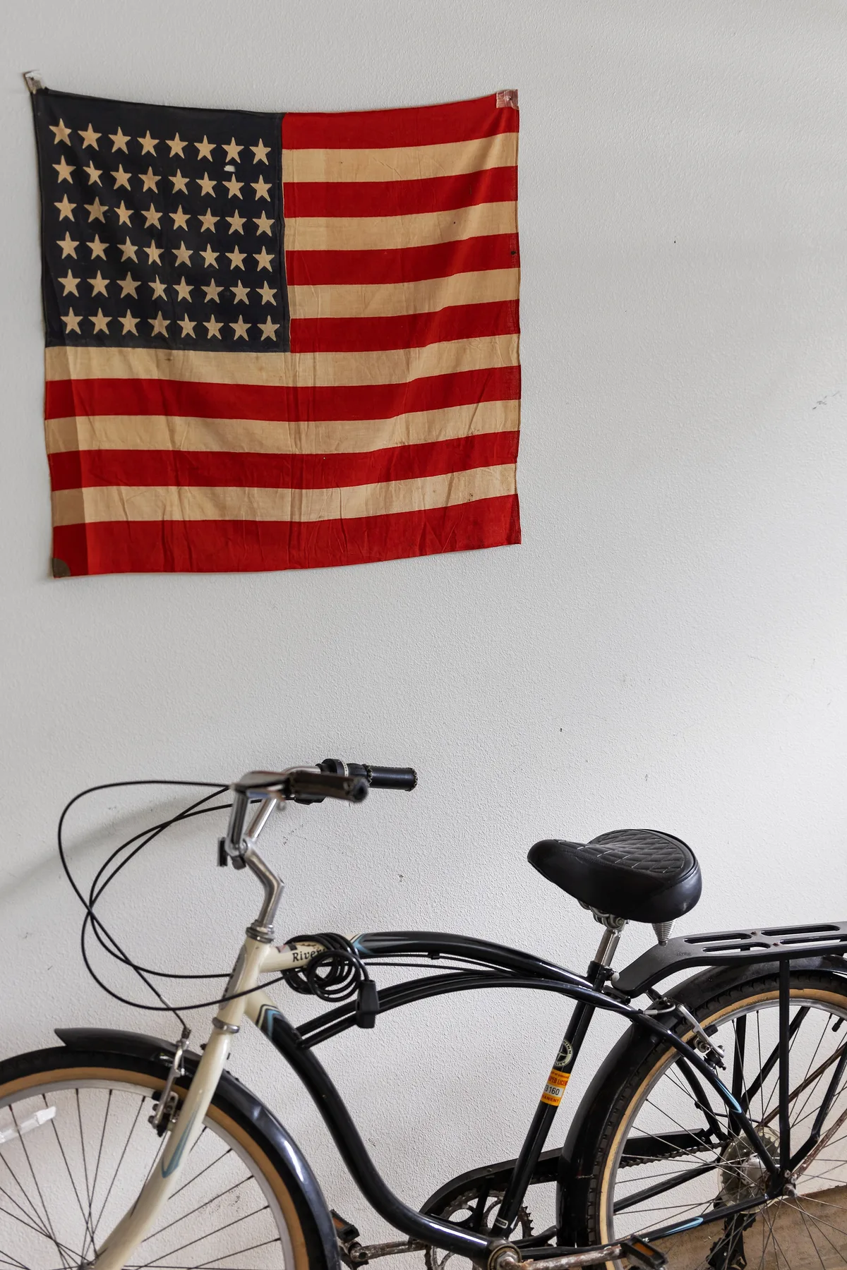 A bicycle stands against a white wall beneath a hanging American flag.