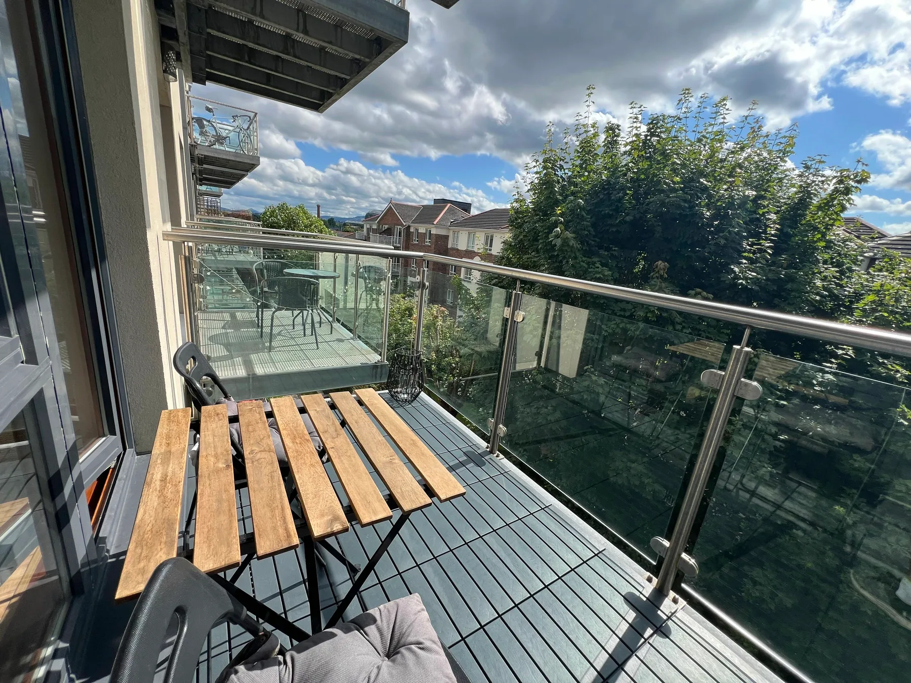 A balcony with a slatted table and glass railing overlooks trees and nearby houses.