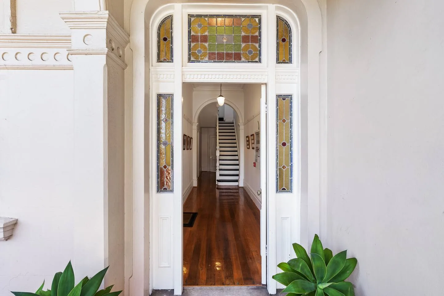 Una entrada ornamentada da paso a un pasillo con suelo de madera, vidrieras y una escalera.