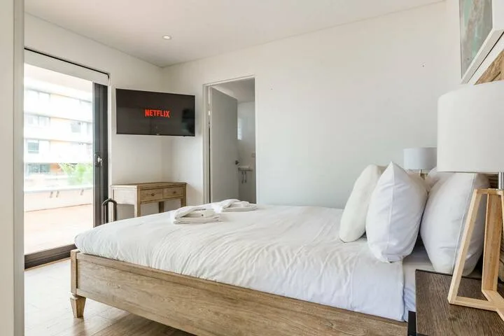 Un dormitorio luminoso con una cama bien tendida y un televisor en la pared.