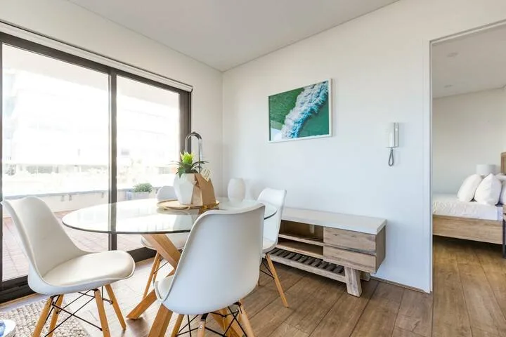 Un comedor luminoso tiene una mesa redonda junto a grandes ventanas y una obra en la pared.