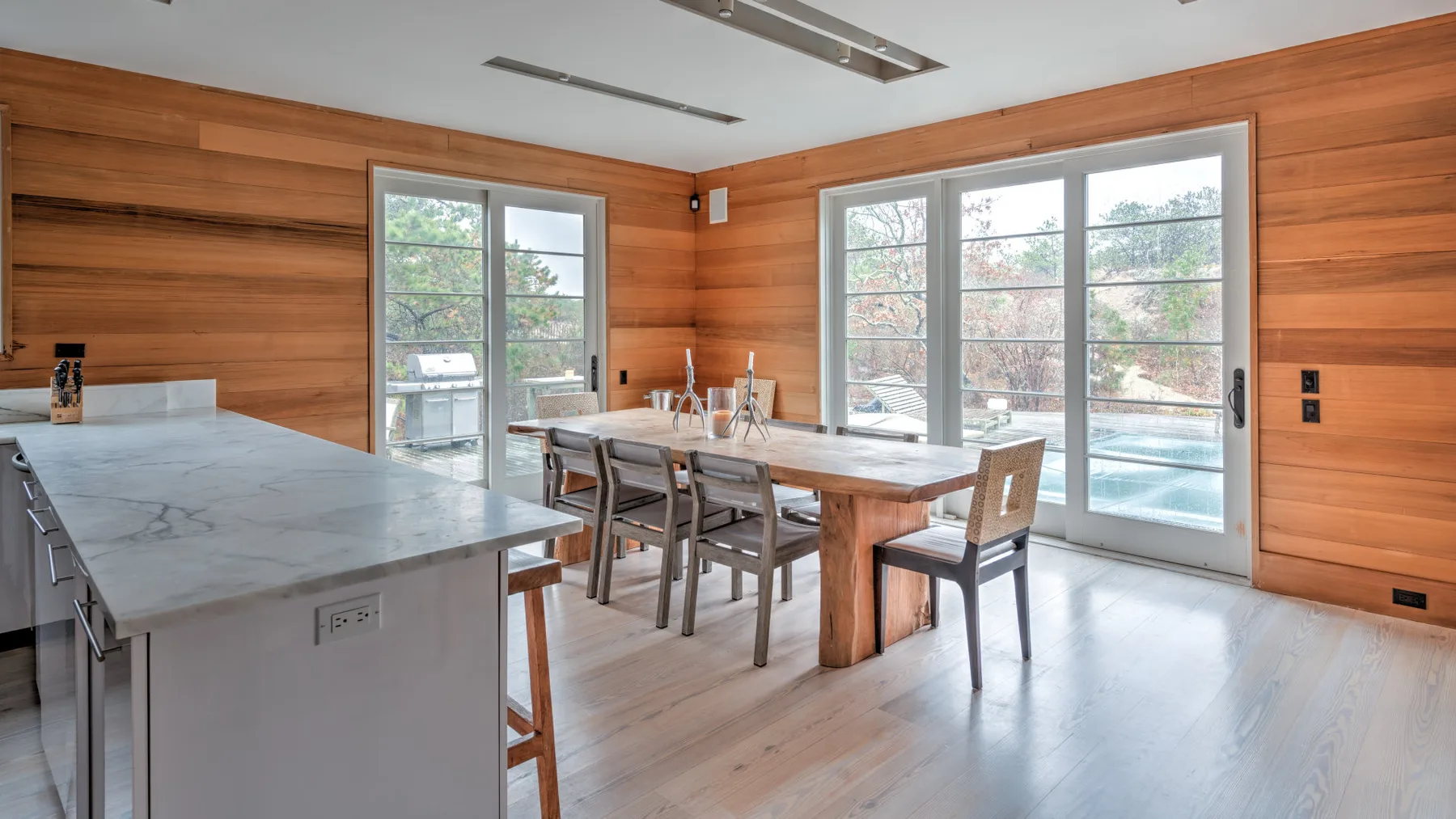 Un comedor con una mesa de madera y grandes ventanas con vista a una piscina exterior.