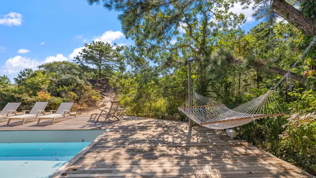 Una terraza de madera junto a una piscina, con tumbonas y una hamaca entre los árboles.