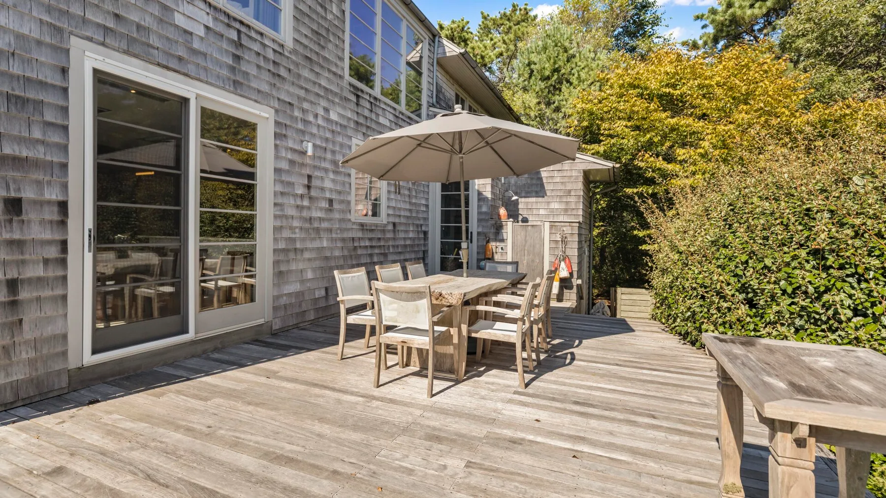 Una zona de comedor al aire libre está en una terraza de madera junto a la casa y un seto.