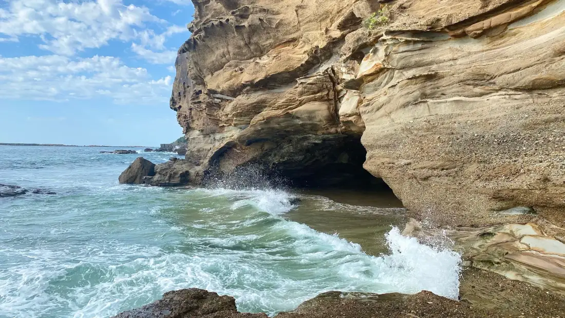 Ocean waves break against a rocky shoreline beneath a large coastal cliff.