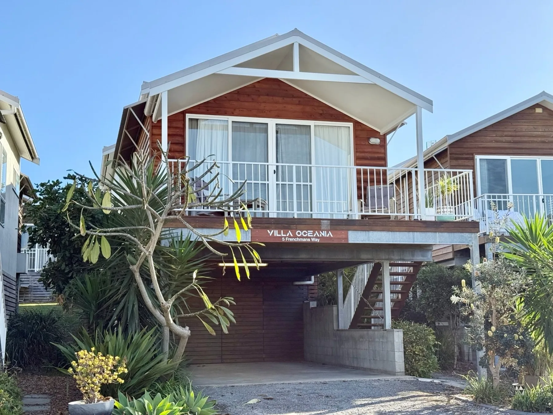 A raised timber beach house has a white balcony and a broad gabled roof.