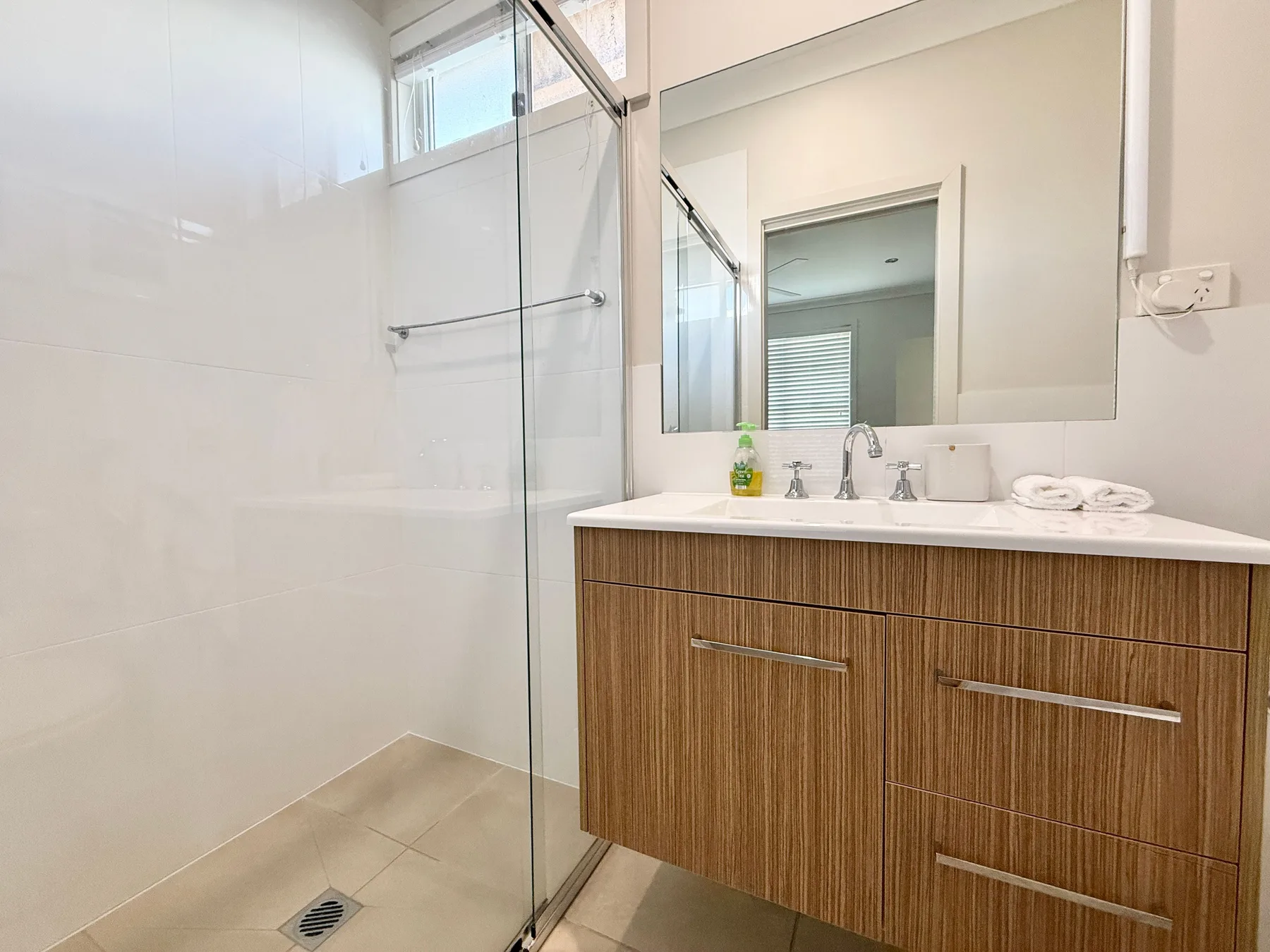 A bright bathroom has a glass shower beside a wide vanity and mirror.