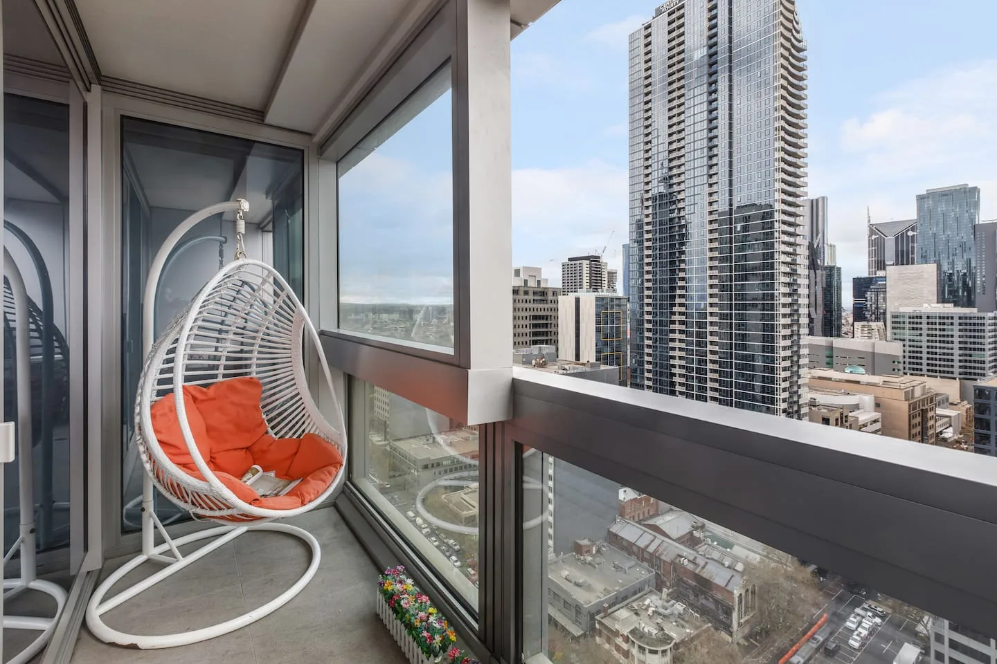 A hanging chair sits on a glass-enclosed balcony overlooking city buildings.