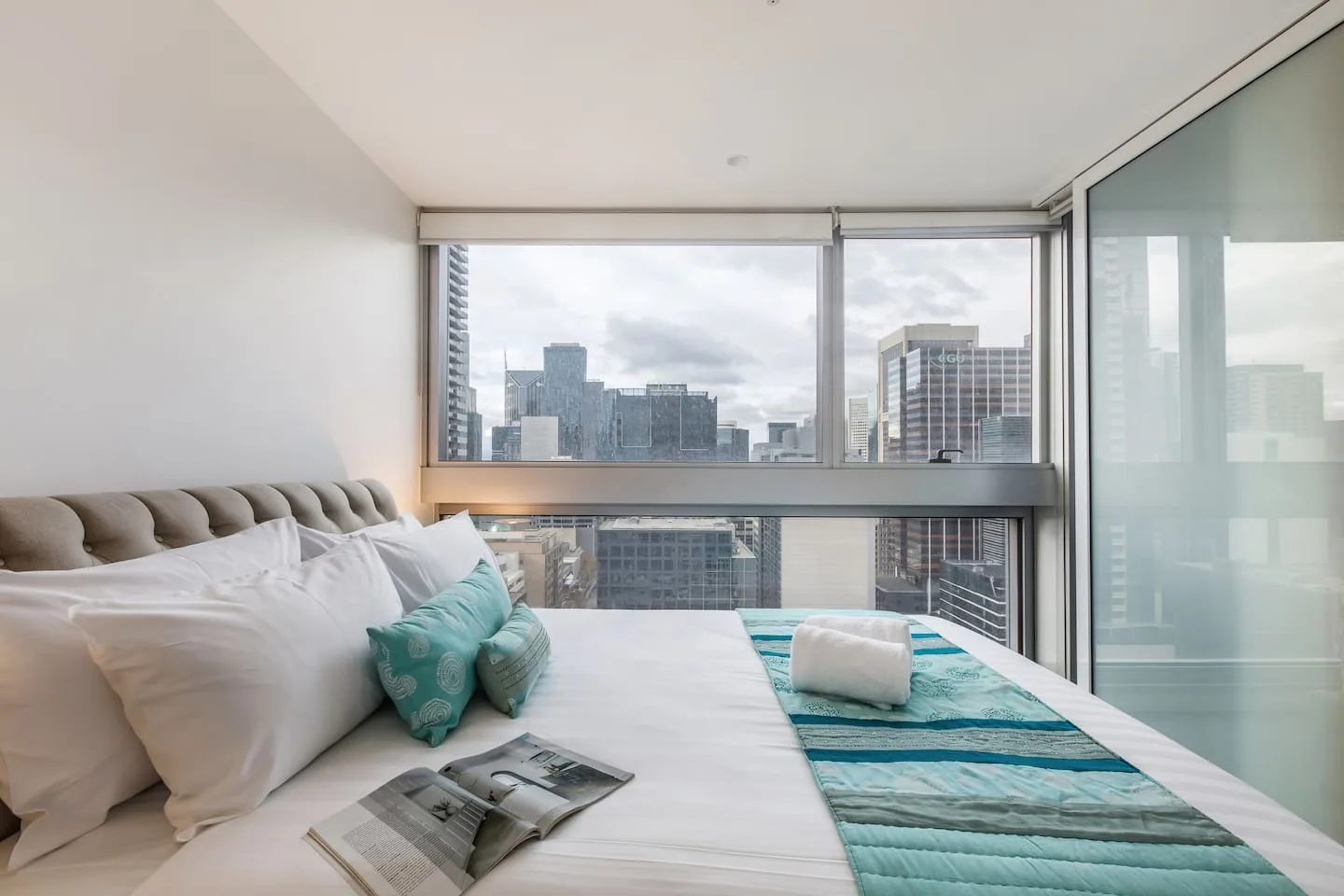 A bedroom with large windows overlooks nearby city buildings.