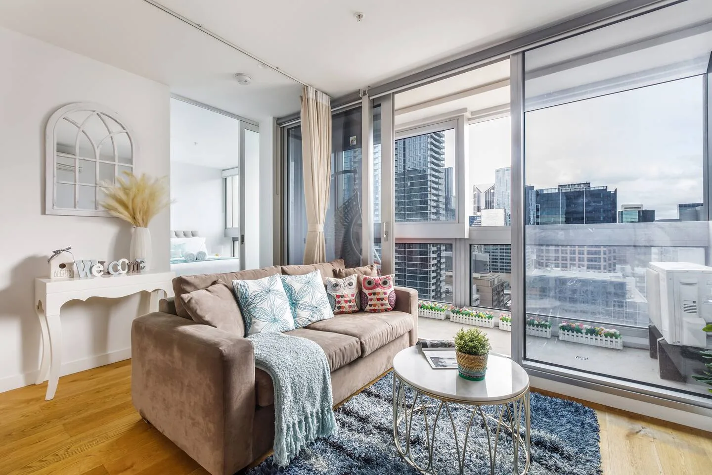 A bright living room with a sofa beside floor-to-ceiling windows overlooking city buildings.