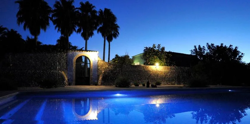 An outdoor swimming pool glows blue beside a garden wall at dusk.