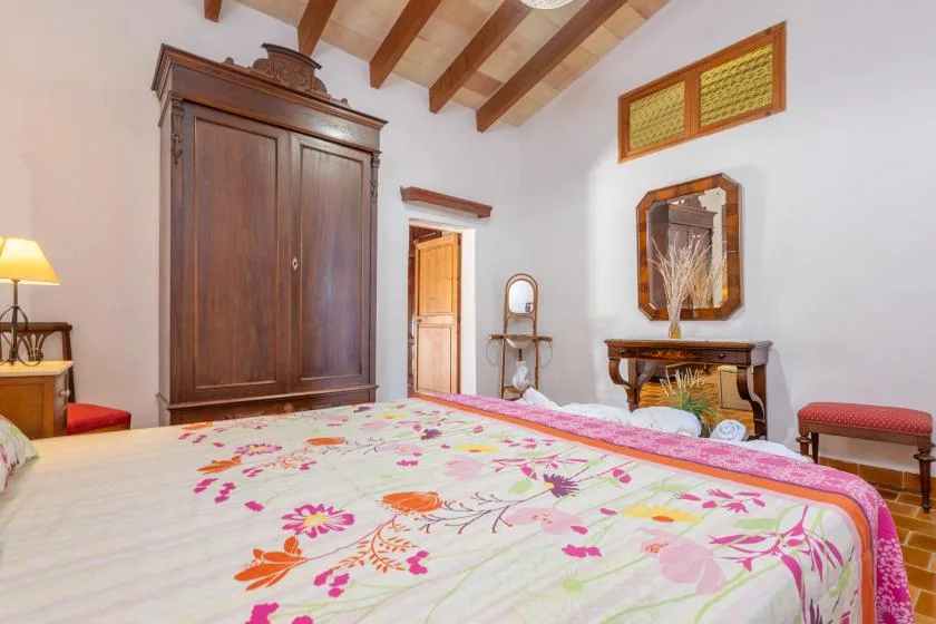 A bedroom with a floral bedspread, wooden wardrobe, and exposed ceiling beams.