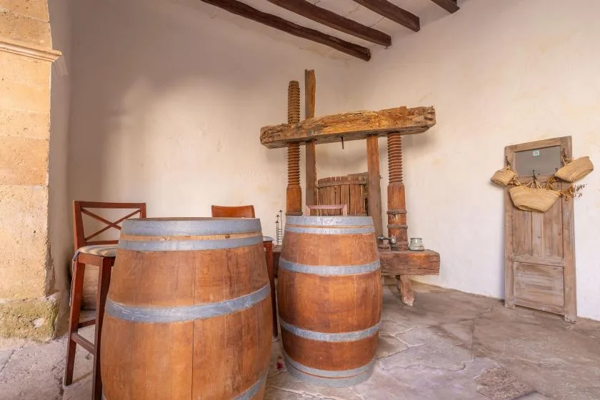 A rustic wine press stands behind two large wooden barrels in a stone-floored room.