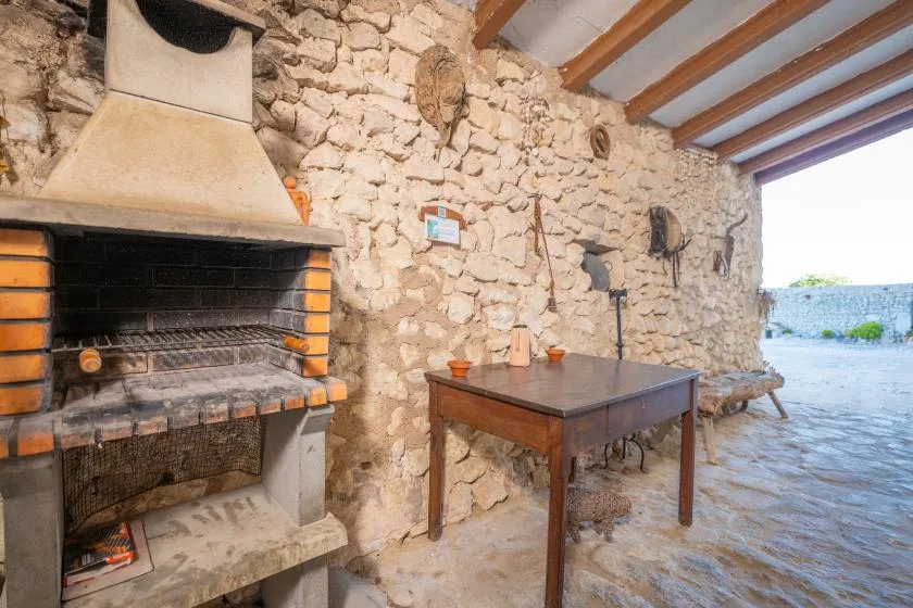 A rustic stone outdoor cooking area with a large built-in grill and a wooden table.