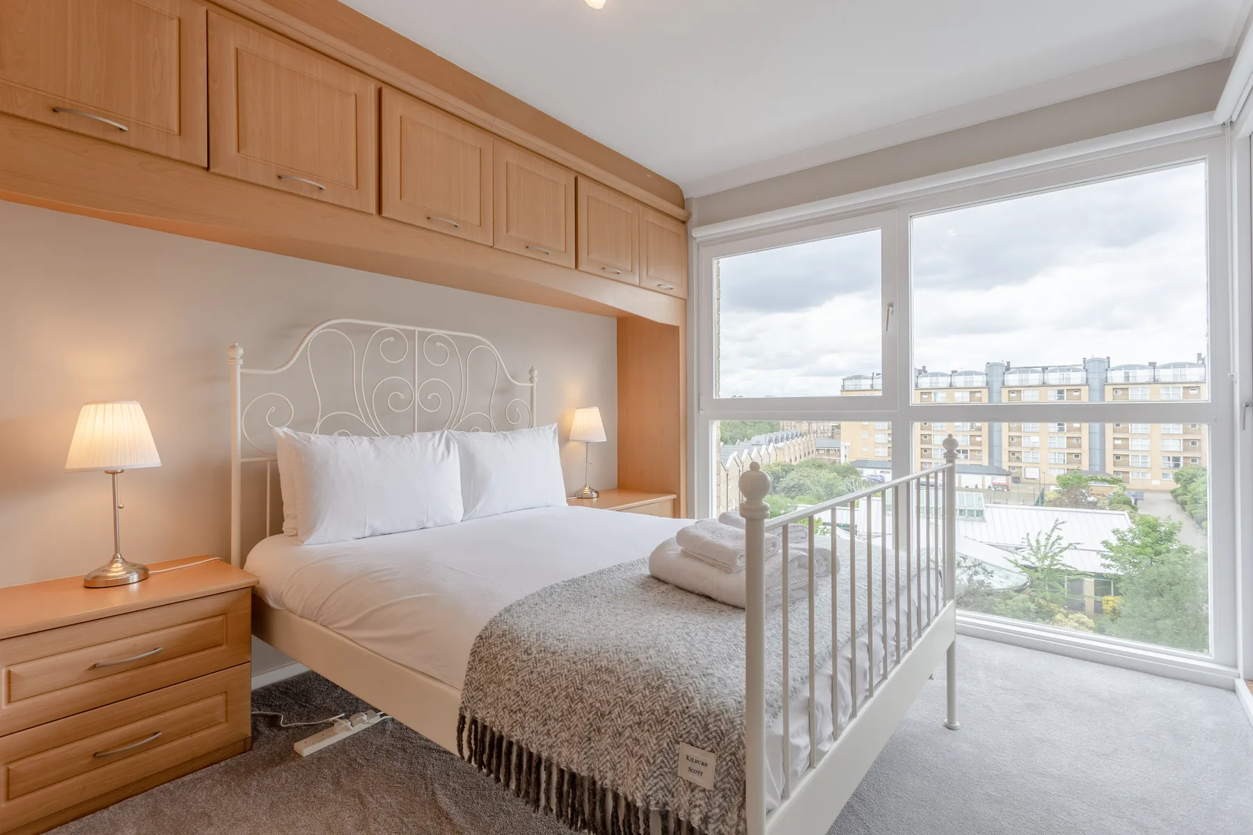 A bedroom with a bed beside large windows overlooking the surrounding buildings.