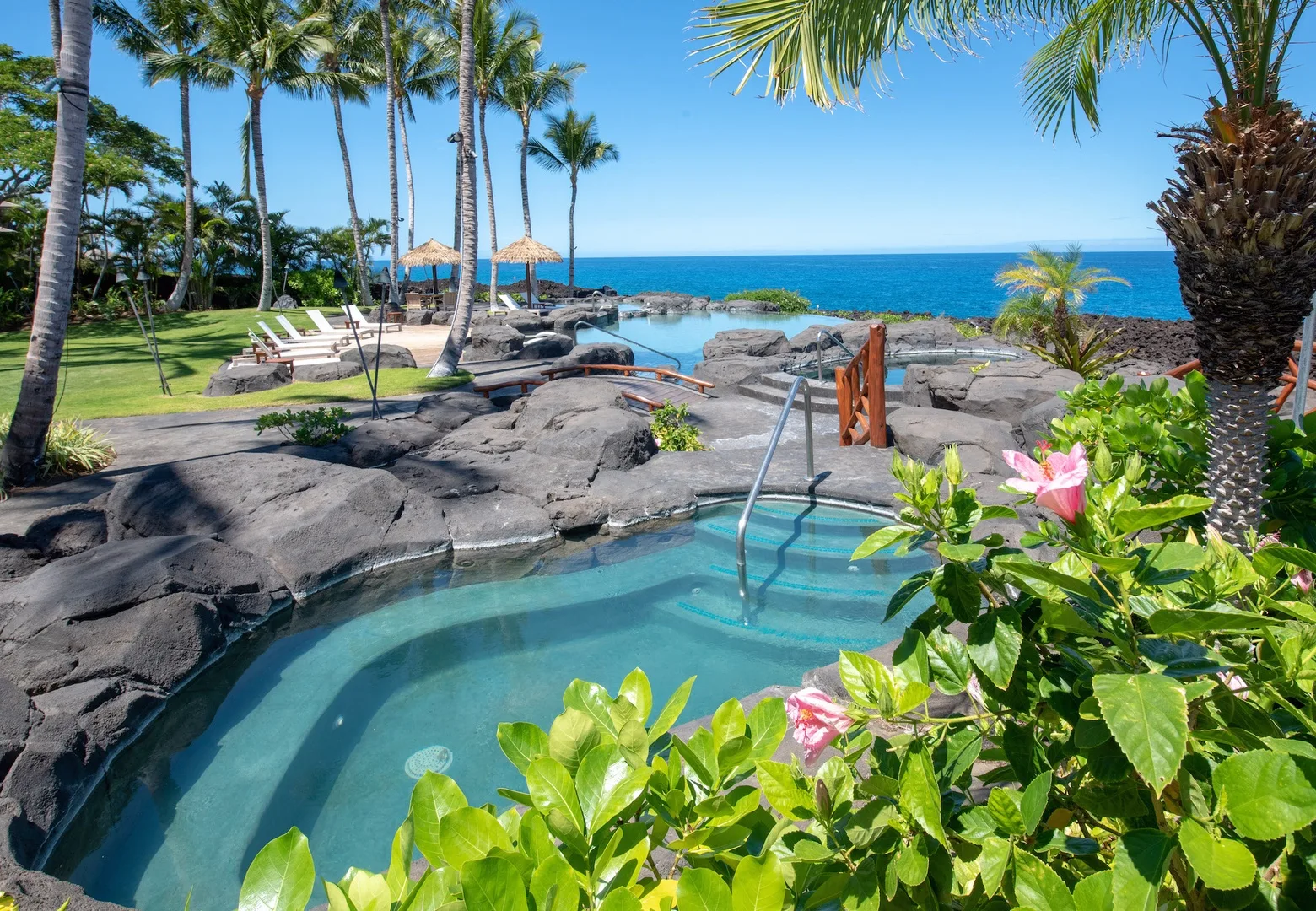 A small outdoor pool sits among rocks and tropical plants beside the ocean.
