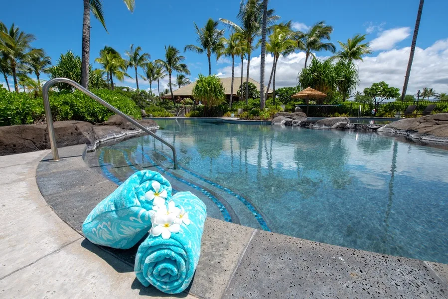 A tropical outdoor swimming pool sits among palm trees, with rolled towels beside the water.