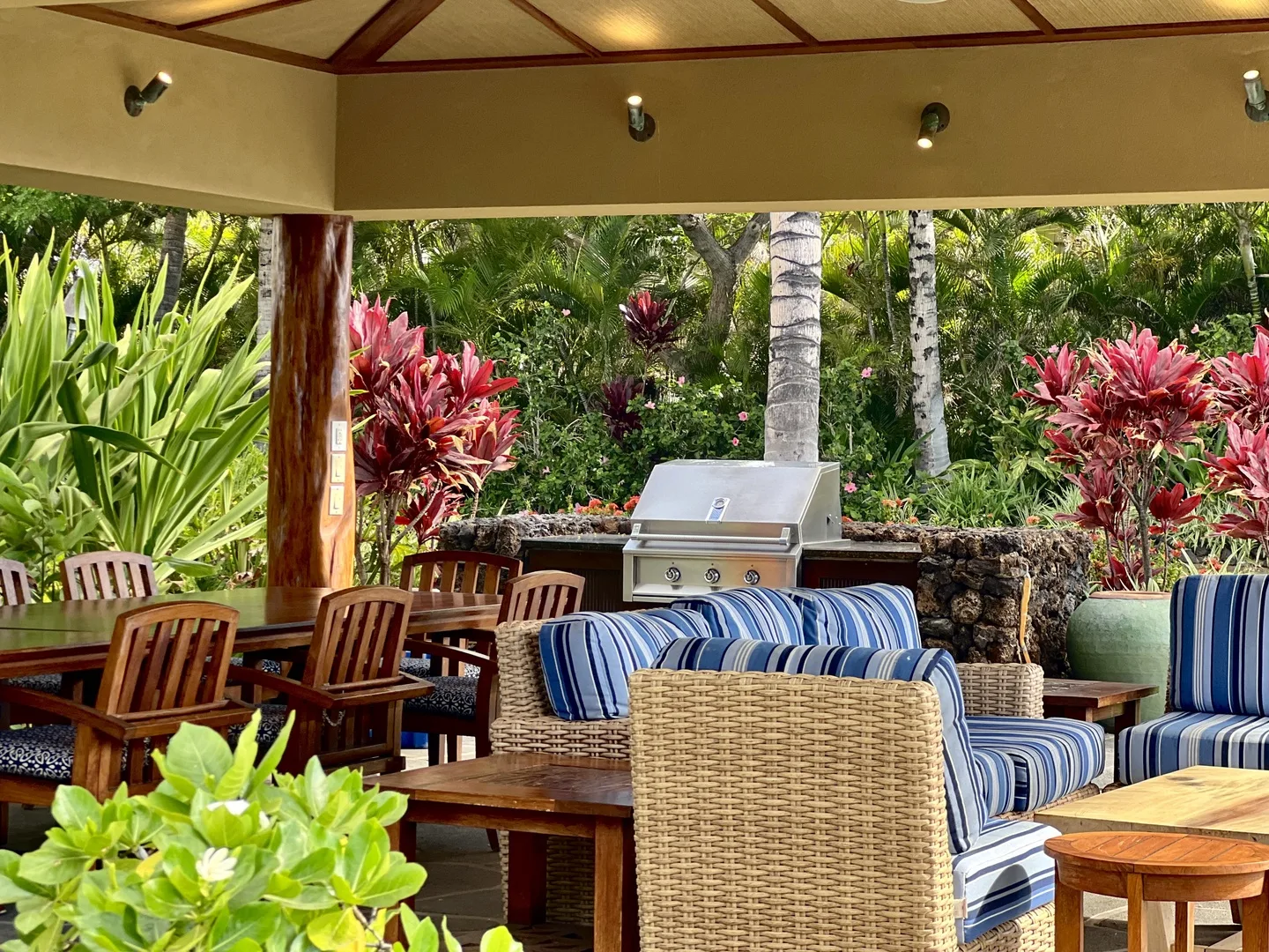 An outdoor patio with cushioned seating and a grill surrounded by tropical plants.