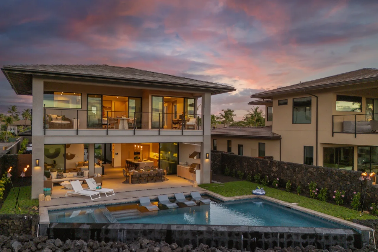 A modern two-story house overlooks a swimming pool beneath a colorful evening sky.