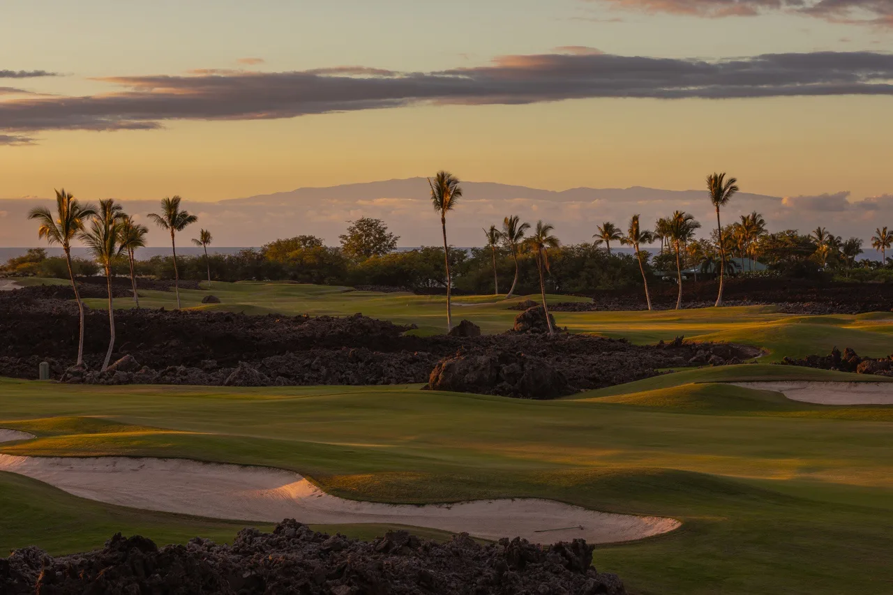 A golf course at sunset, with palm trees and distant mountains.