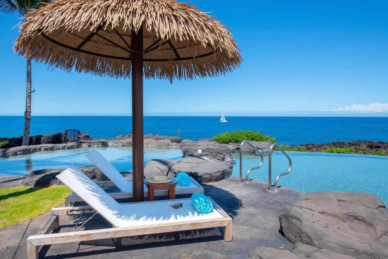 A poolside lounge area overlooks the ocean, with a thatched umbrella shading a chaise lounge.