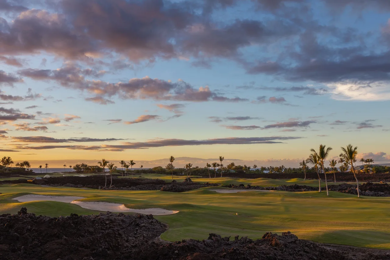 A golf course at sunset, with palm trees and dark rocky ground.