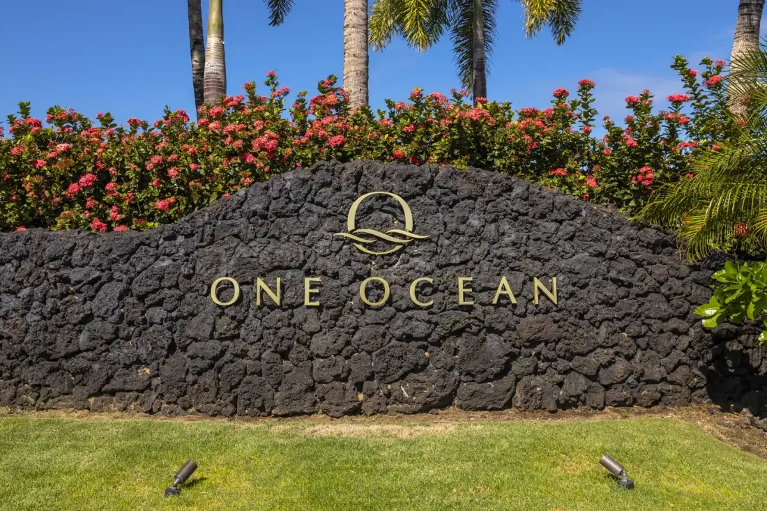 A stone entrance sign stands beside a lawn and flowering shrubs.