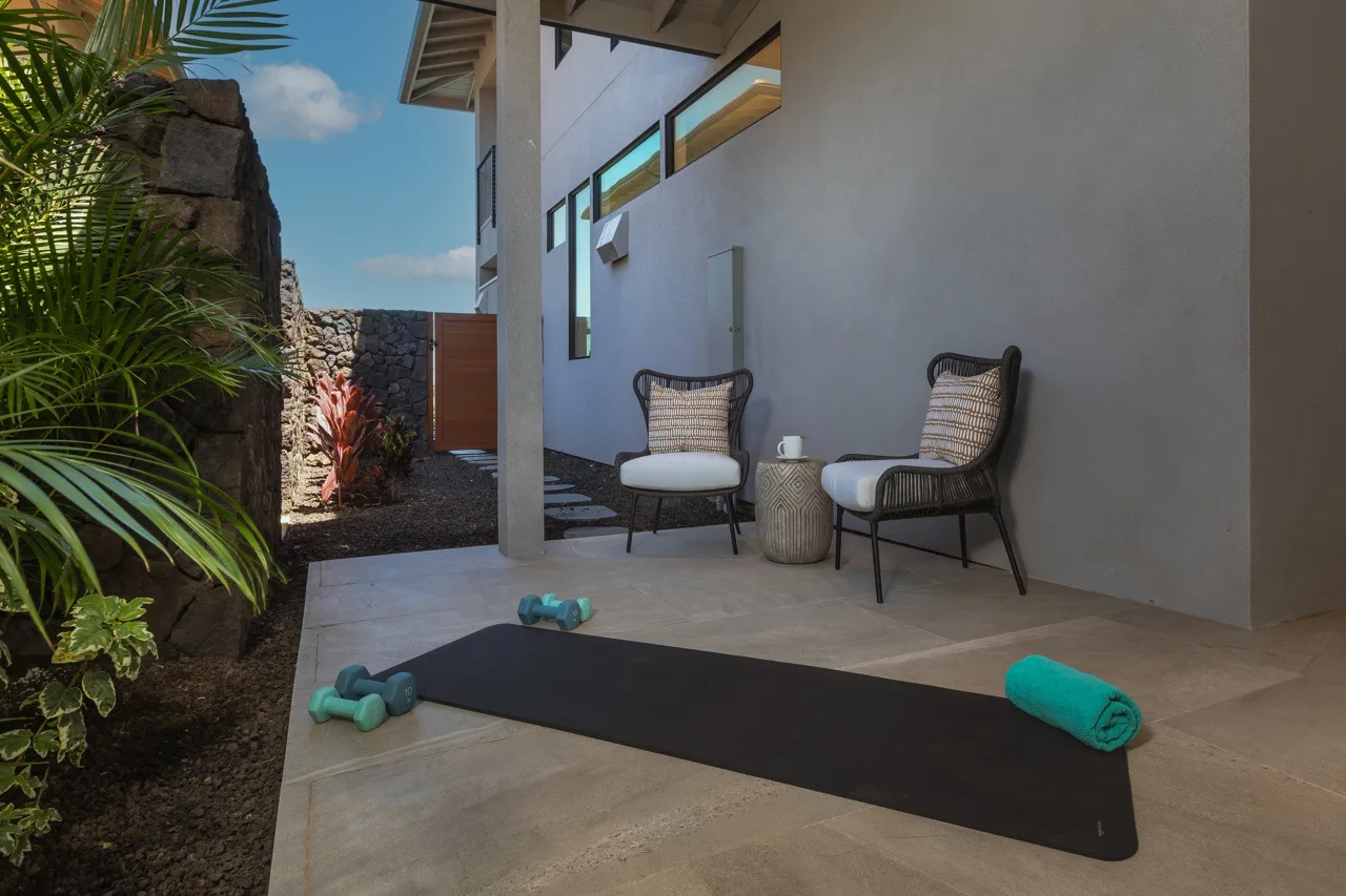 A covered patio has two seats beside a yoga mat and tropical plants.