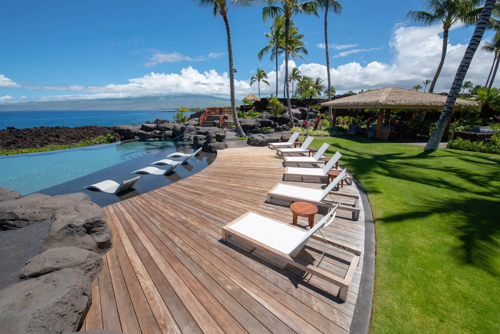 A poolside deck has lounge seating, palm trees, and a grassy lawn.