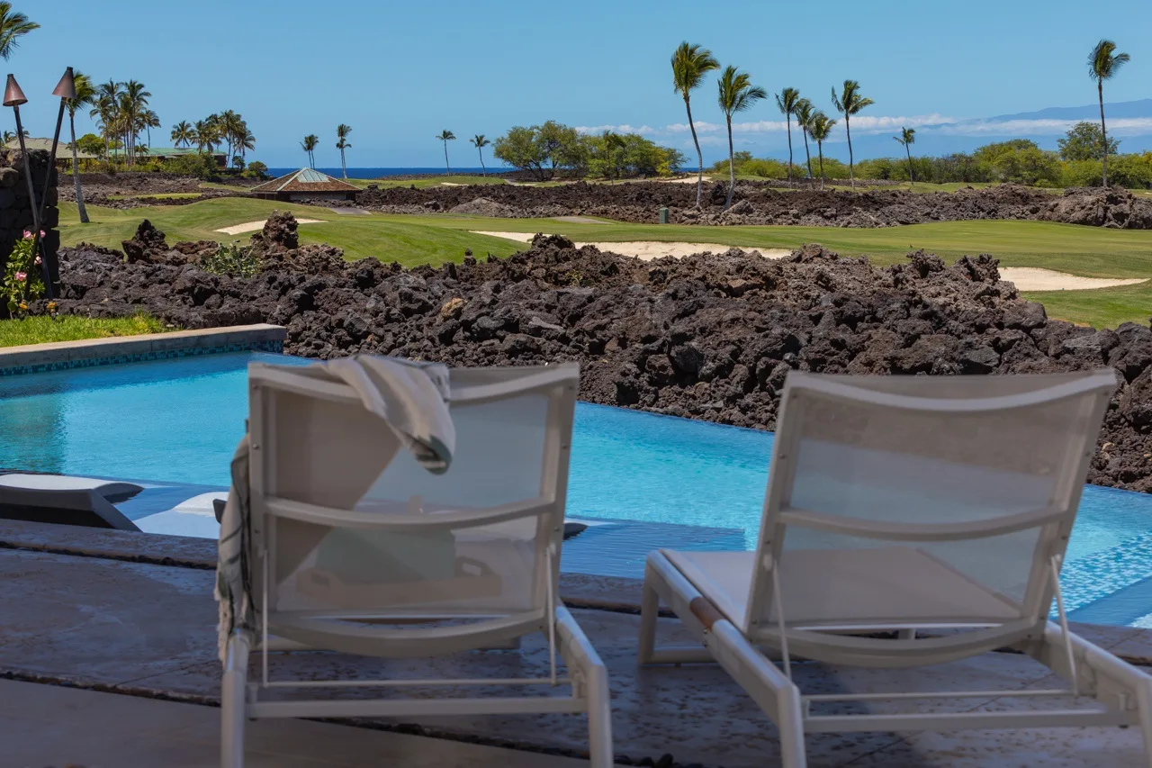 Poolside seating overlooks a rocky landscape, green grounds, and palm trees.