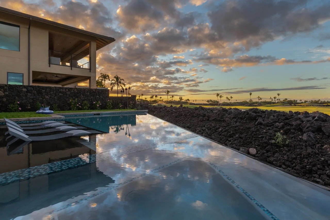 An outdoor pool beside a modern building reflects a cloudy sunset sky.