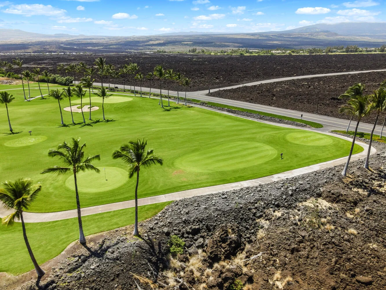 Un campo de golf verde con palmeras se encuentra junto a un amplio paisaje volcánico.