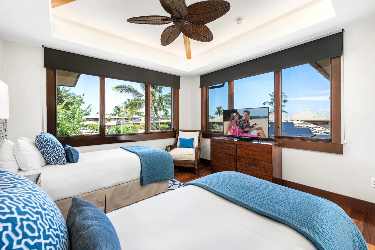 Un dormitorio luminoso con dos camas, ropa de cama azul y grandes ventanas con vistas a la vegetación.