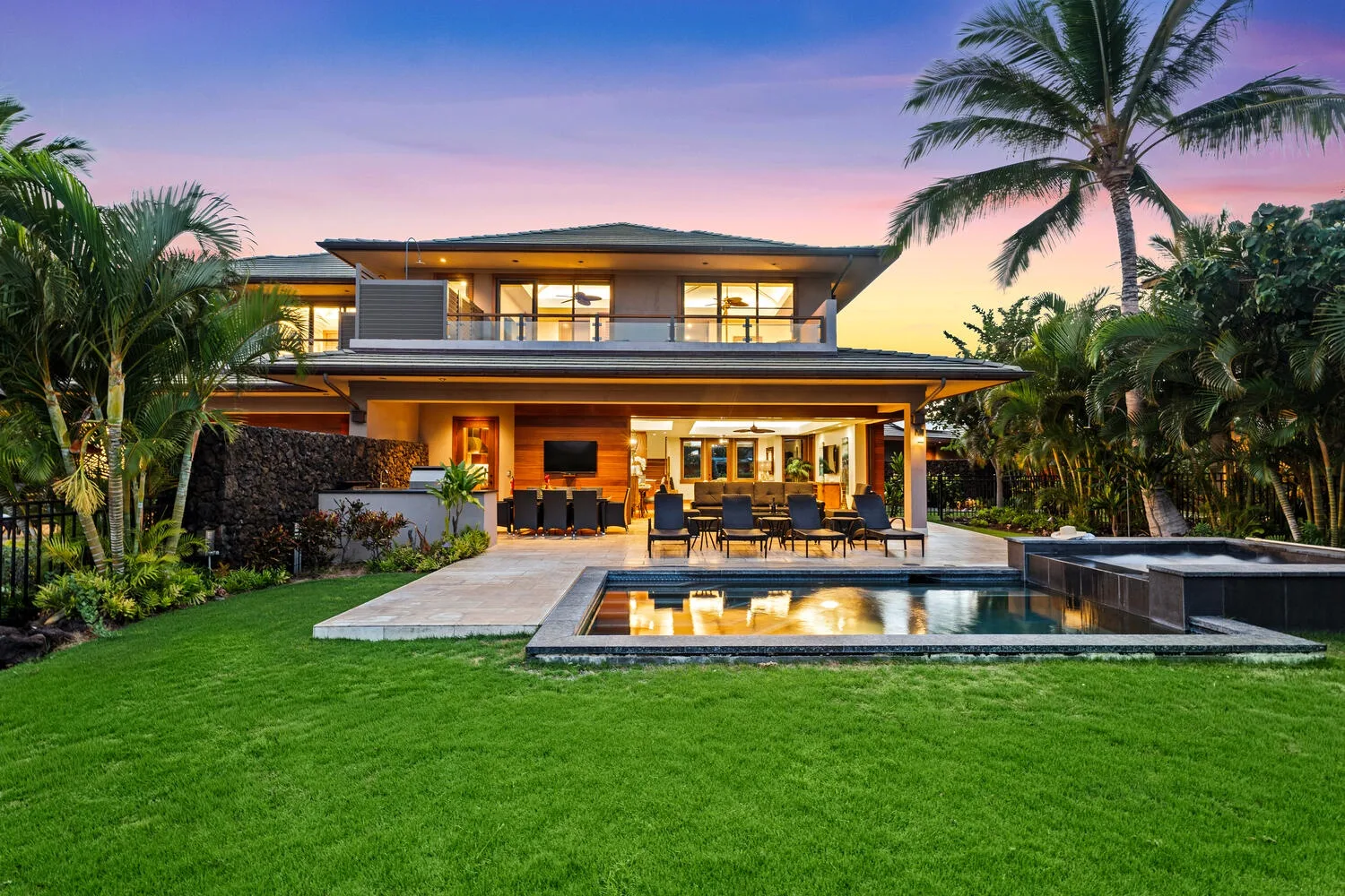 A modern house opens onto a lawn, patio, and illuminated swimming pool at dusk.