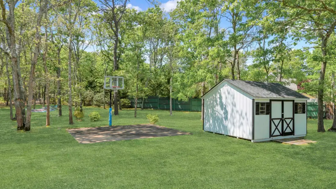 Un patio arbolado y cubierto de césped cuenta con un cobertizo y una canasta de baloncesto.