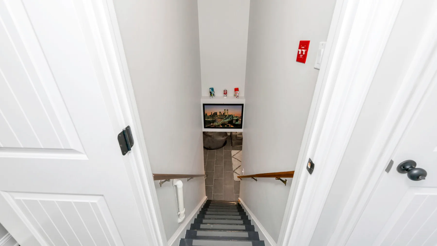 Una escalera interior estrecha desciende entre paredes blancas, con un pasamanos de madera y un cuadro.