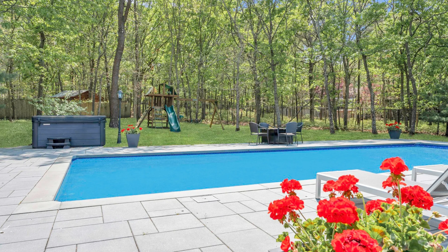 Una piscina al aire libre está rodeada por un patio pavimentado, flores rojas y árboles.