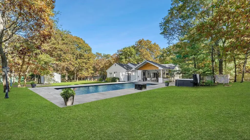 Una casa y una piscina se encuentran más allá de un amplio césped verde, bajo árboles maduros.