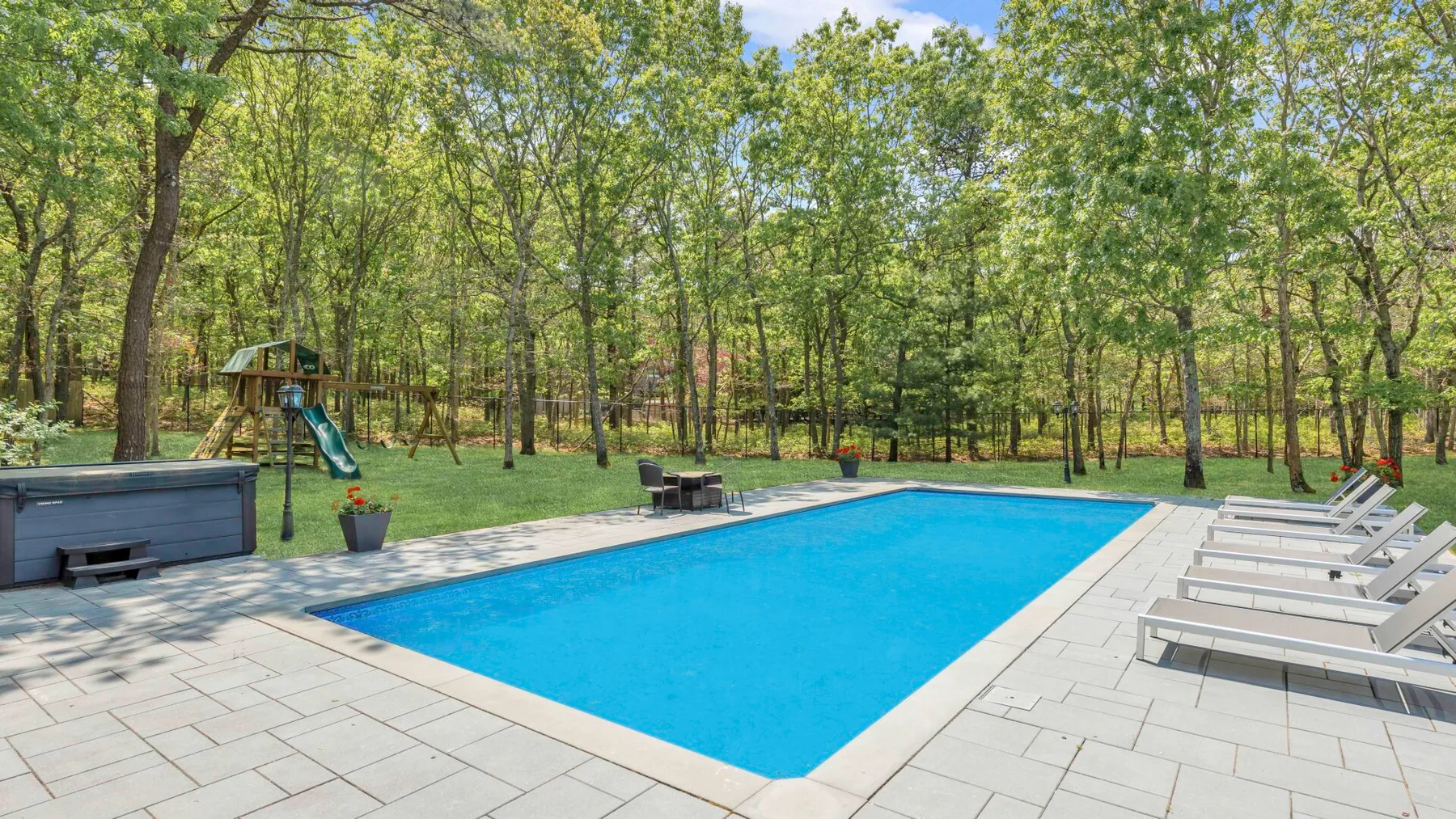 Una piscina al aire libre está rodeada por una terraza pavimentada y árboles.
