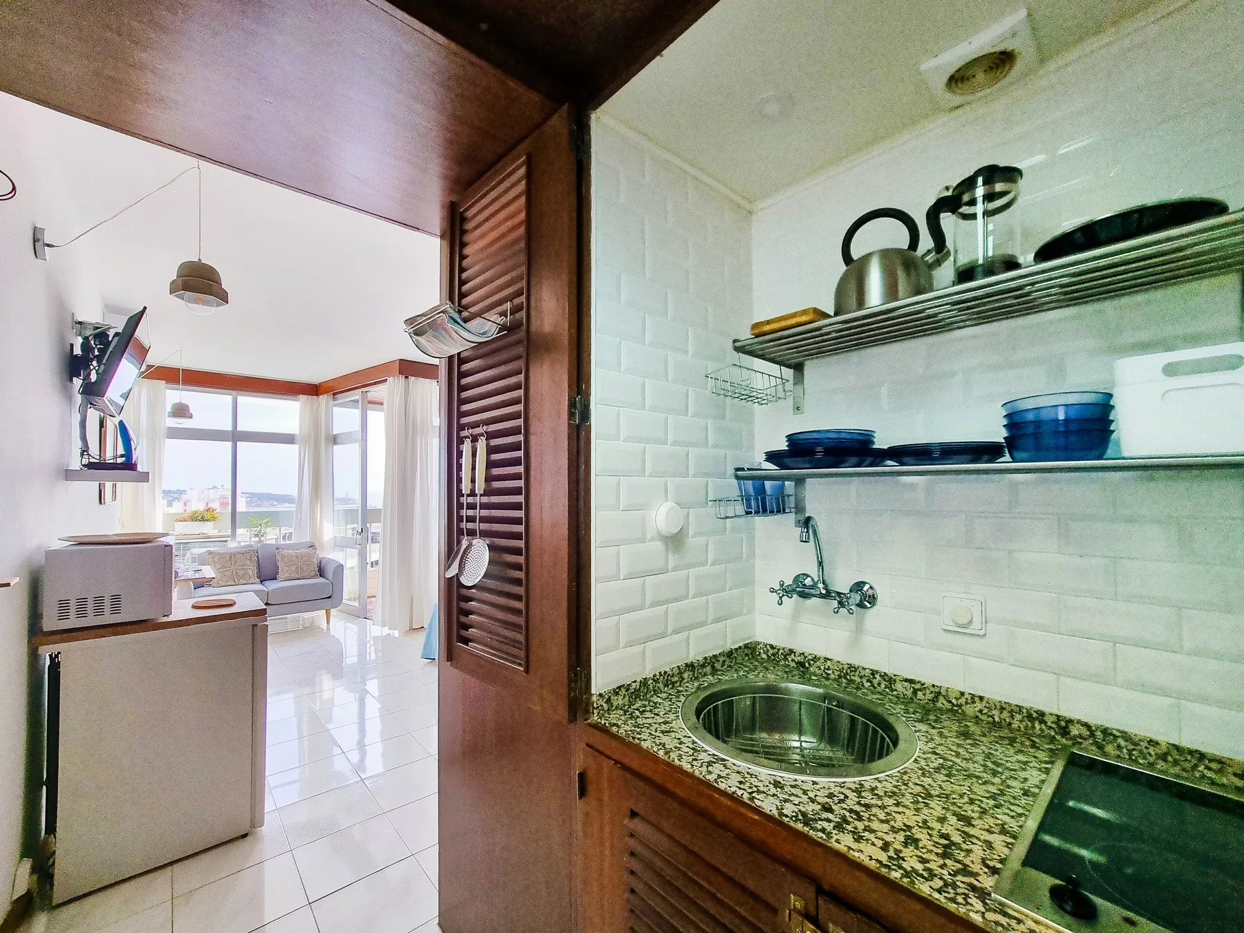 Una cocina compacta con fregadero y estantes abiertos junto a una sala luminosa.