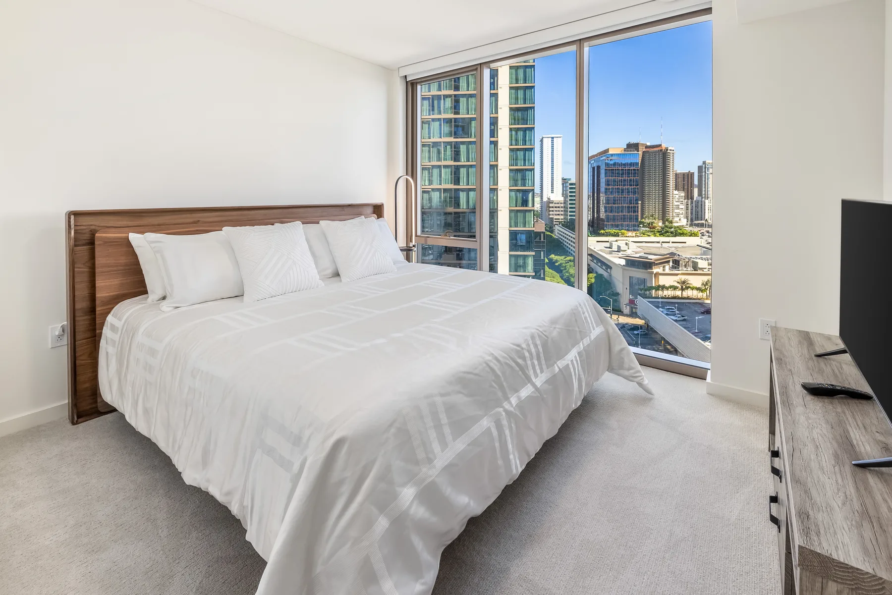 A bright bedroom with a neatly made bed beside large windows overlooking city buildings.