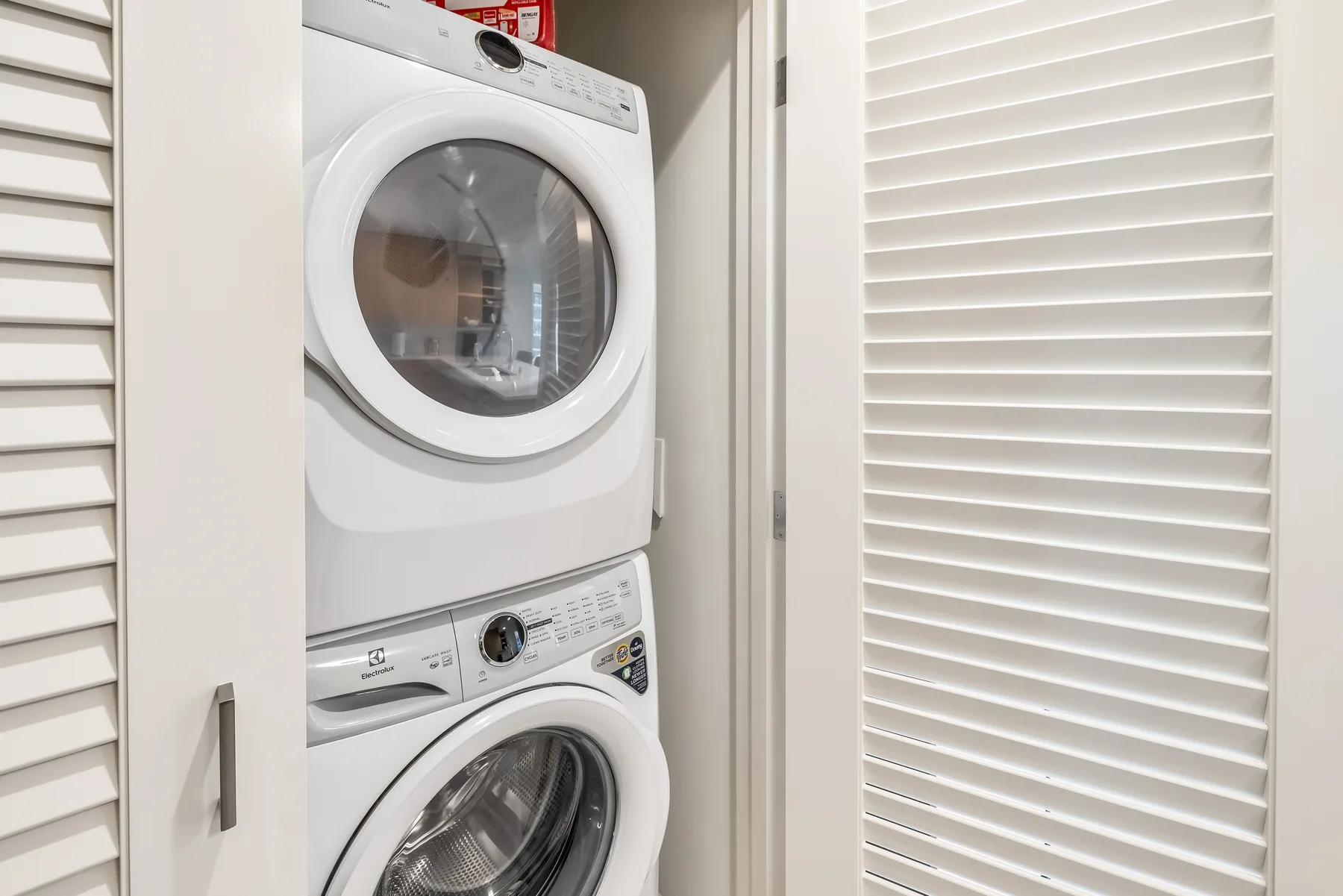 A laundry area with a stacked washer and dryer beside white louvered doors.