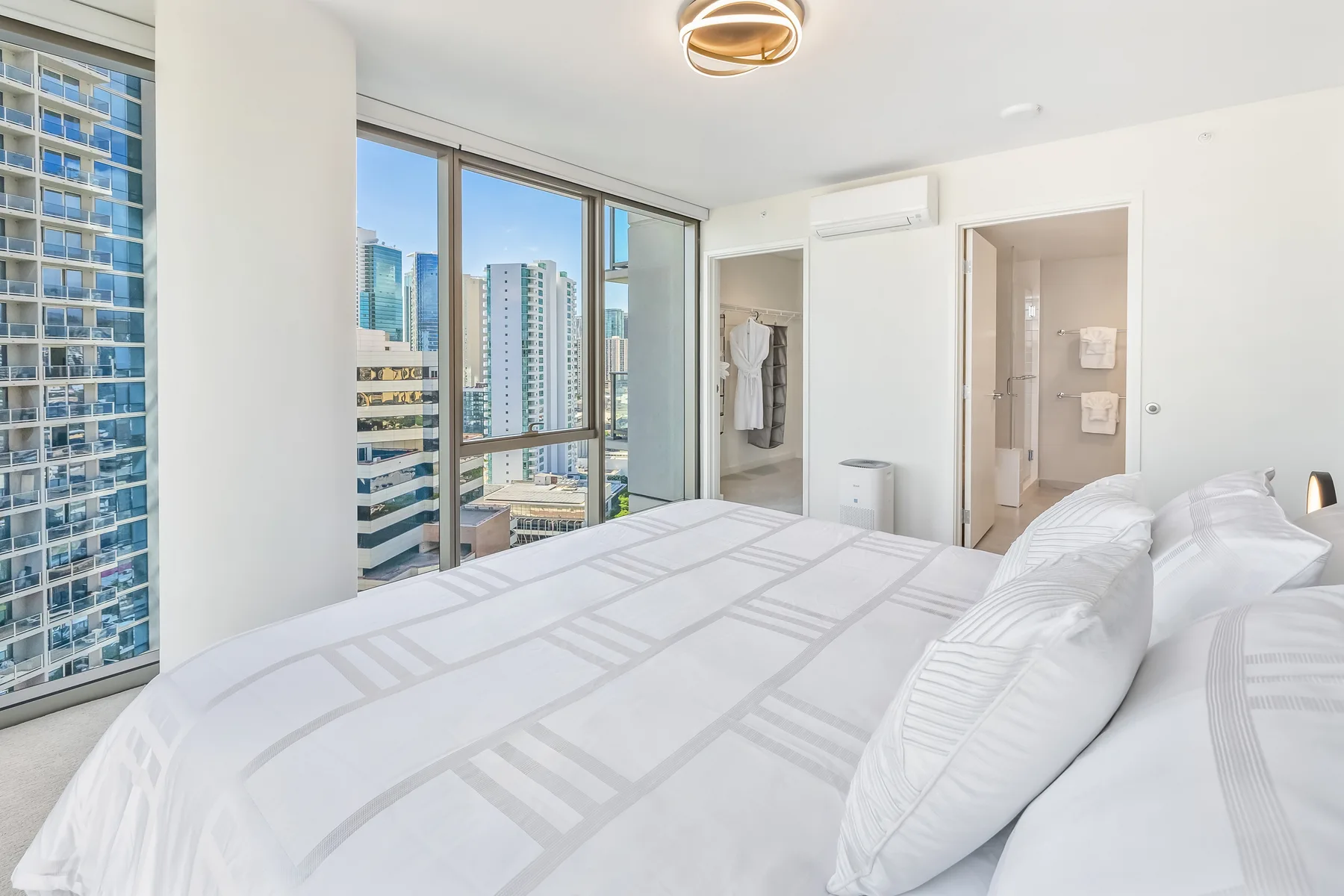 A bright bedroom with a neatly made bed and large windows overlooking city buildings.