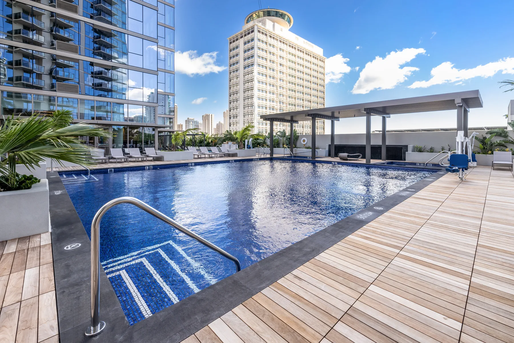 A rooftop swimming pool sits beside a wooden deck and modern buildings.
