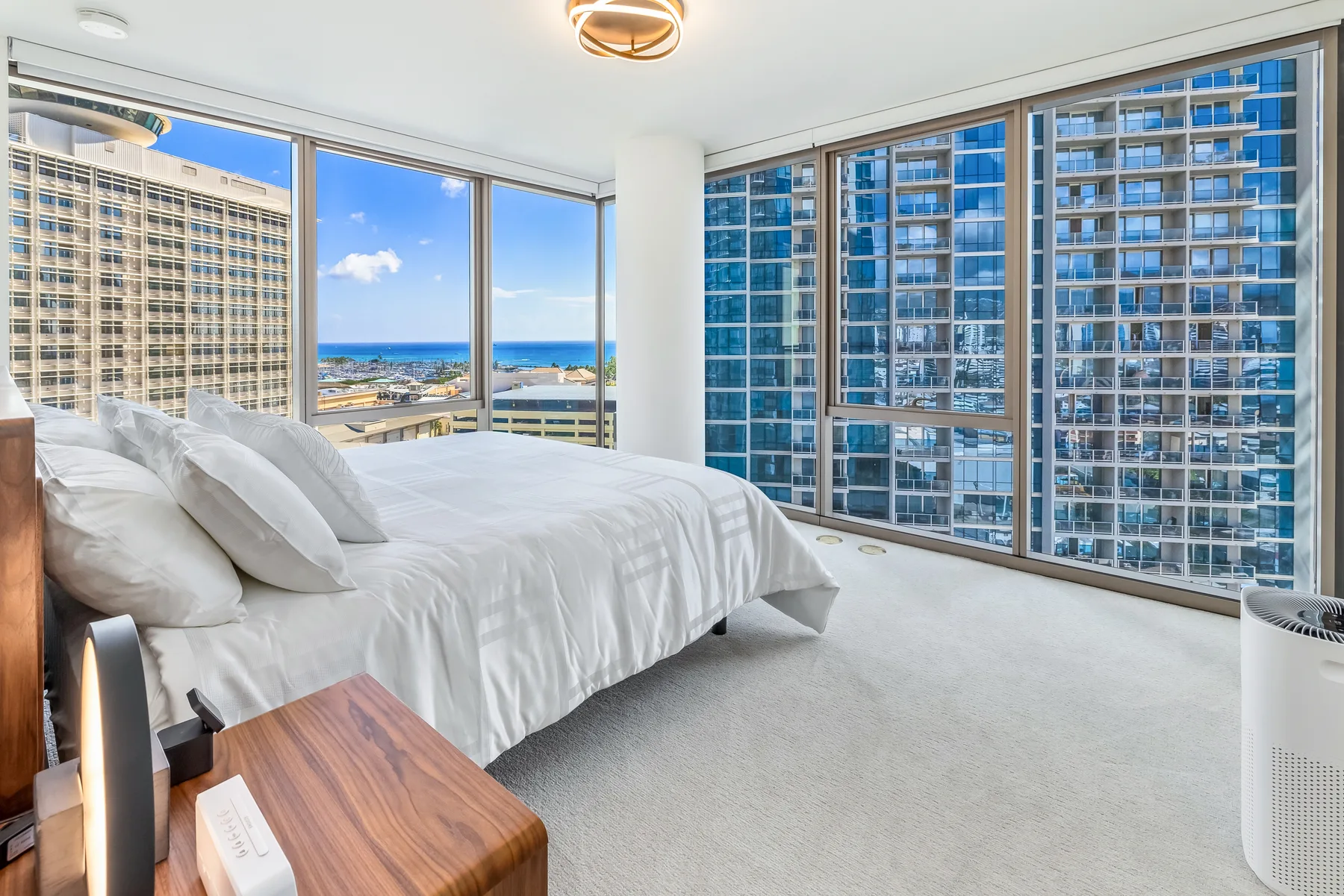 A bright bedroom with a bed beside large windows overlooking nearby buildings and the coast.