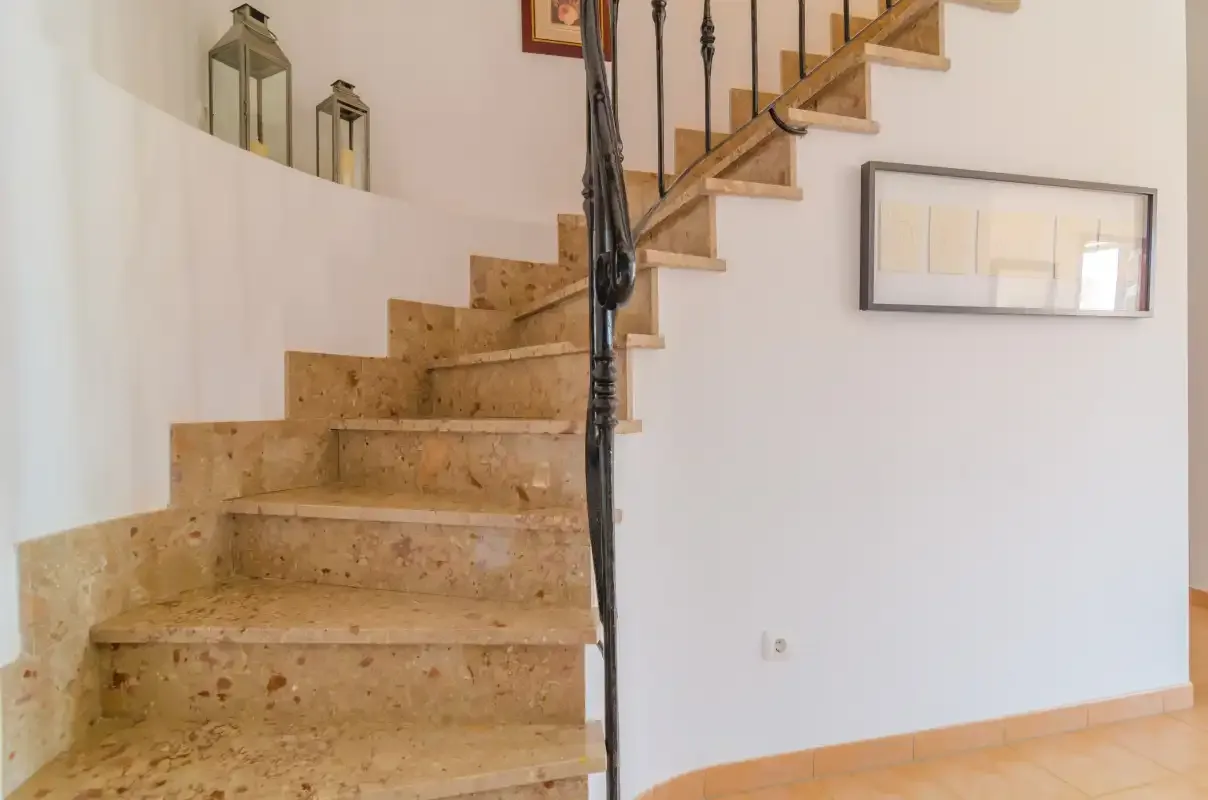 A staircase with stone steps and a black metal railing rises beside a white wall.