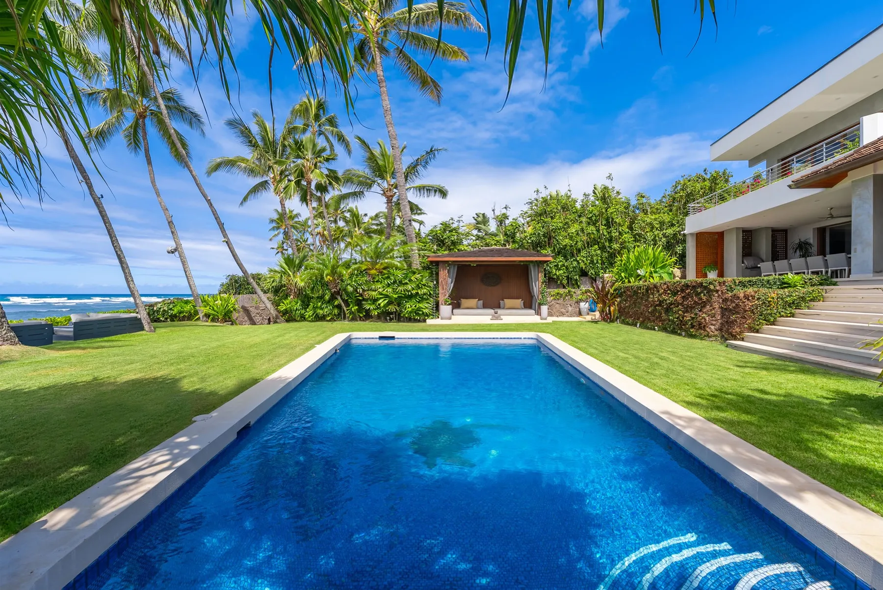 An outdoor swimming pool sits in a tropical garden beside a modern building.