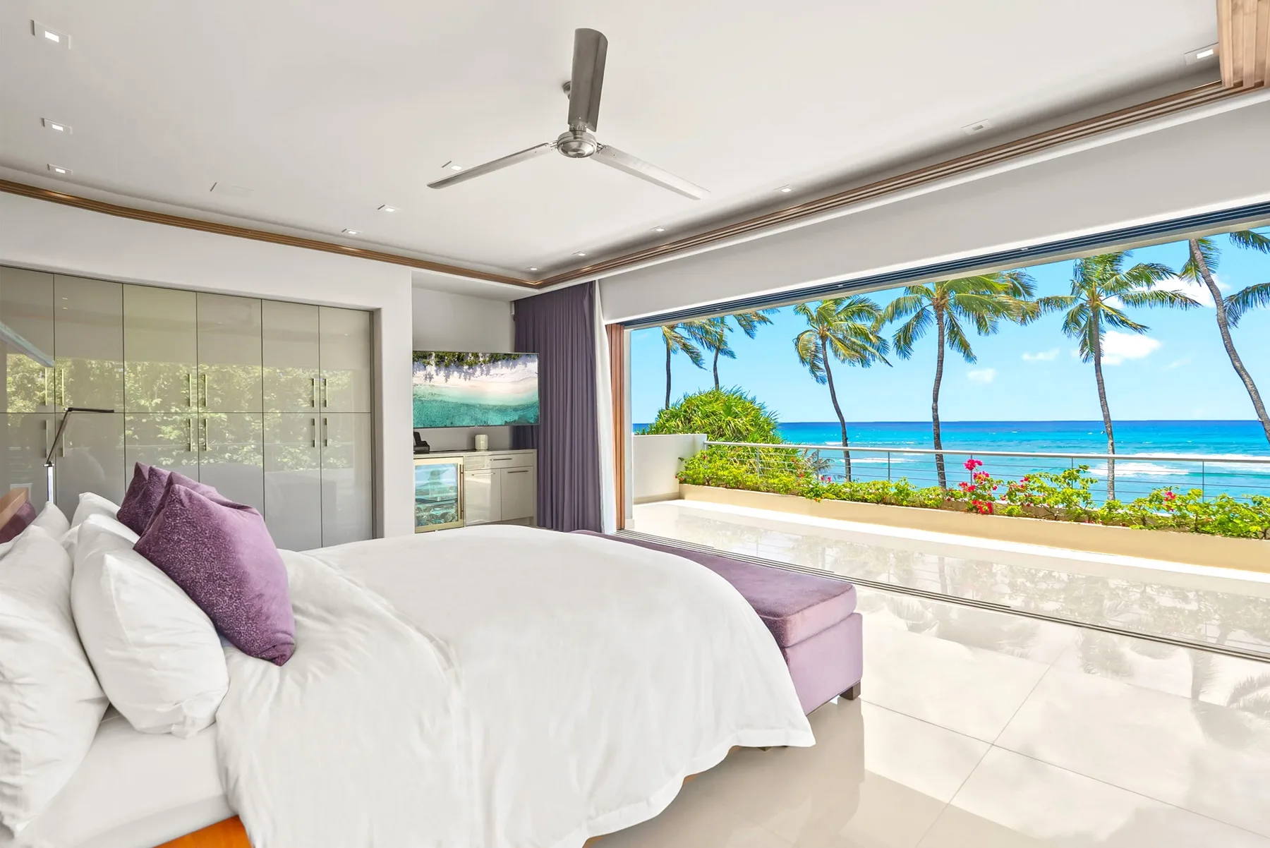 A bedroom with white bedding and a wide window overlooking palm trees and the ocean.