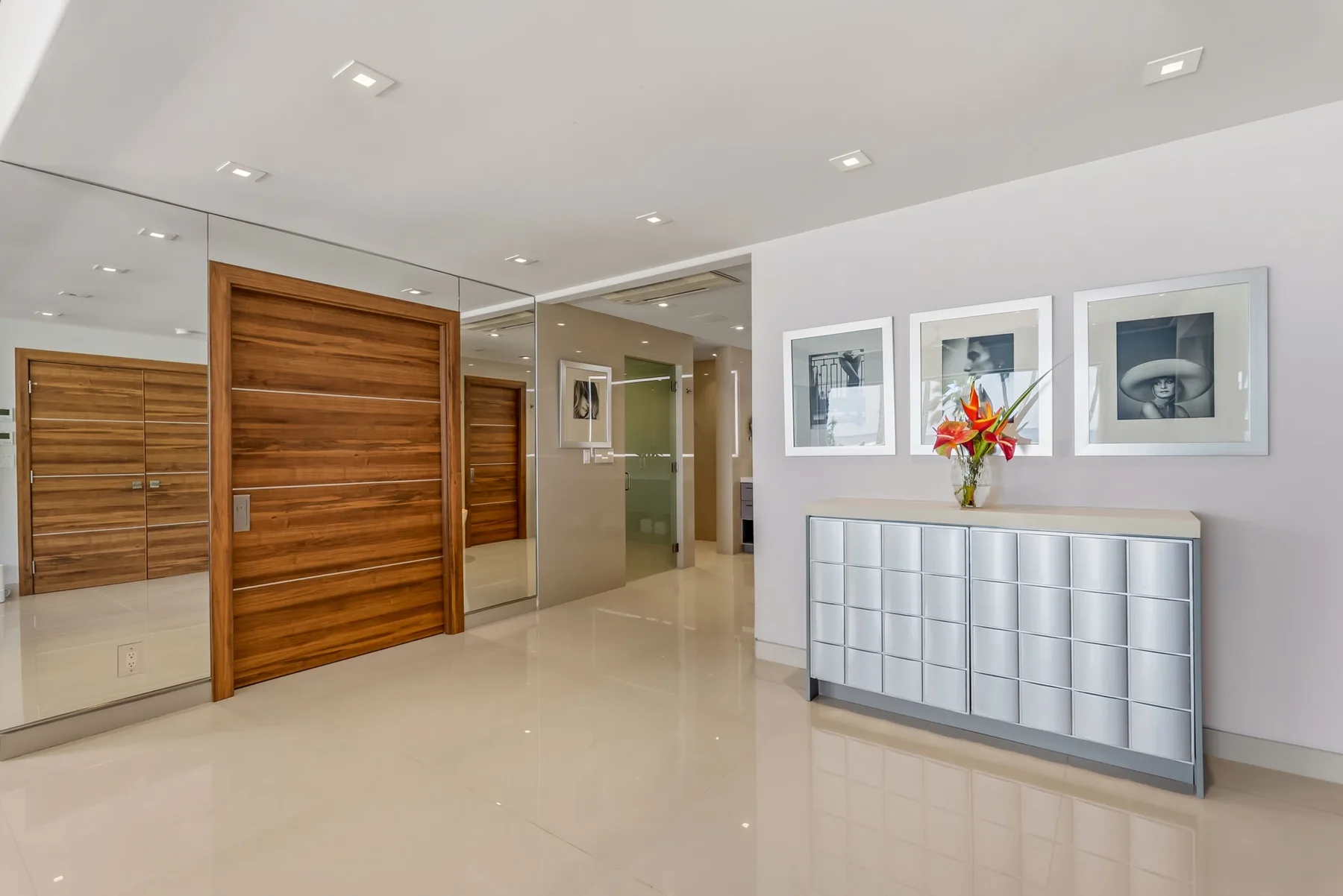 A modern hallway with mirrored walls, wooden doors, and a silver cabinet.